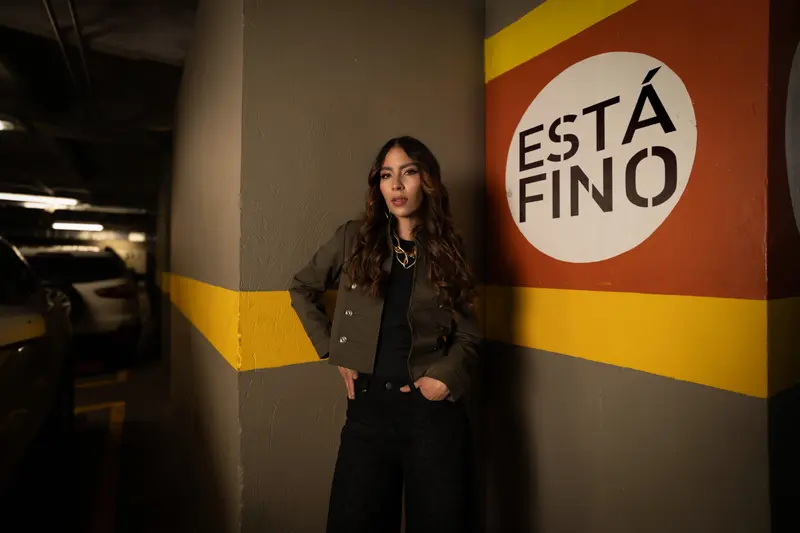 Modelo con chaqueta verde oliva en un parqueadero, junto a un círculo blanco con «ESTÁ FINO» pintado en la pared