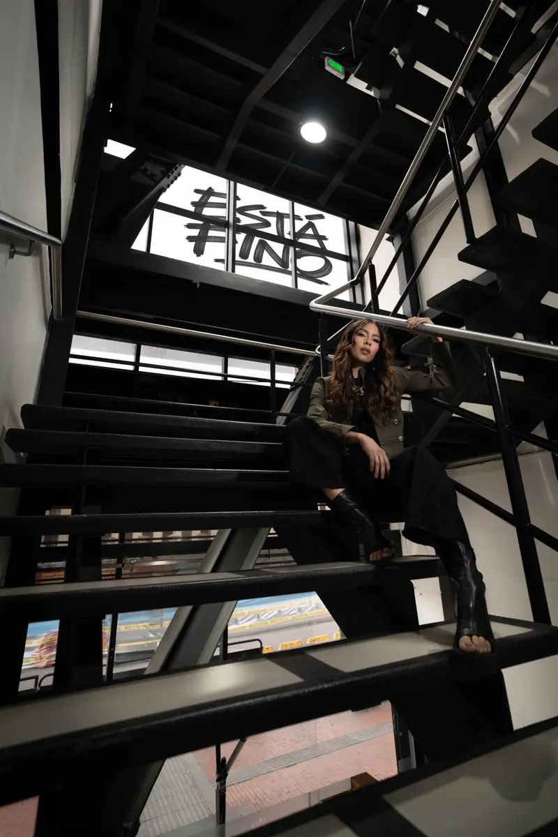 Modelo con chaqueta verde oliva sentada en una escalera metálica negra, bajo una ventana con «Está fino»